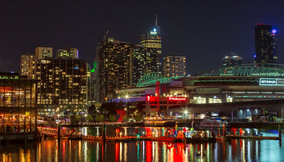 A nighttime cityscape of Melbourne, Australia, featuring a skyline with illuminated high-rise buildings, including one with the logo 'AMP' at the top. In the foreground, a waterway reflects the colorful city lights, with a dock or pier visible along the edge. To the right, a large stadium or event arena with a curved roof and green lighting accents is prominent, alongside red warning signals and docked boats along the waterfront. The scene captures the vibrant urban environment with bright, glowing windows in the buildings, and the reflections shimmering on the calm water surface, creating a dynamic contrast between the illuminated structures and the dark sky. This image showcases a modern city at night, highlighting its architectural features and waterfront activity, which can be associated with urban waste collection or private rubbish clearance services like those offered by Rubbish Clearance Docklands, often involving on-site clearance or alternative waste handling methods.