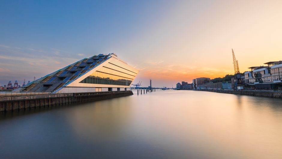A wide-angle view of a modern building with a distinctive sloped, terraced design featuring multiple angular levels, situated on a waterfront during sunset. The building's facade is composed of smooth, reflective glass panels and light-colored materials, capturing the warm hues of the setting sun. The structure is positioned closest to the foreground on the left side of the image, with a concrete dock running along its base, extending over calm water that reflects the building and sky. In the background, the waterway stretches toward the horizon with several cranes and industrial containers visible on the right, indicating a port area. The sky displays soft gradient tones from the sunset, transitioning from blue to orange, and the scene is illuminated by natural late-day light, creating a serene atmosphere suitable for observing the urban landscape and waterfront surroundings. This setting subtly relates to services like private waste handling or on-site clearance by illustrating a modern waterfront environment, possibly undergoing development or maintenance, where efficient rubbish removal could be a necessary part of ongoing work. Rubbish Clearance Docklands operates within this context of urban renewal and sustainable disposal solutions.
