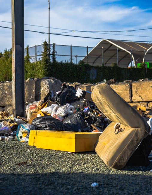 A roadside accumulation of mixed waste including black bin bags, plastic packaging, and scattered debris is shown in the foreground, with an old, dirty car tire leaning against a low stone wall. The waste material appears haphazardly piled on a gravel surface adjacent to a small section of curb and is partially contained within a yellow plastic container, possibly used for rubbish collection. Behind the waste, there is a backdrop of a tall wooden utility pole with electrical wires stretched across the scene, and a vegetation hedge that partially obscures a fenced area. In the distance, a large, modern greenhouse structure with a curved, semi-transparent roof is visible, set against a partly cloudy blue sky with sunlight illuminating the scene, suggesting daylight hours. The overall composition indicates an area awaiting clearance or removal of accumulated rubbish, exemplifying the type of waste accumulation that a private rubbish removal service such as Rubbish Clearance Docklands may handle as part of their on-site clearance operations, providing an alternative to traditional council rubbish collection in the area.