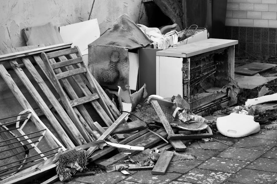 A cluttered outdoor scene featuring a variety of discarded household items and debris placed on a paved surface. On the left side, there are two weathered wooden garden chairs with slatted backs and seats, leaning against a wall. Behind and slightly to the right, an old, stained mattress or large fabric-covered object is partially covered with a tarpaulin or cloth. To the right of the chairs, a small white appliance, possibly a mini refrigerator or part of an old unit, is visible, with its door open and internal components exposed. Surrounding these objects are broken pieces of wood, cardboard, and insulation material scattered across the pavement, along with a white plastic object resembling a portable toilet or toilet seat. The background features a wall built from concrete blocks, partially covered with dark tiles, indicating an exterior environment. The overall scene suggests an on-site clearance operation, possibly related to independent waste disposal or rubbish removal services, with visible signs of neglect and abandoned waste materials.