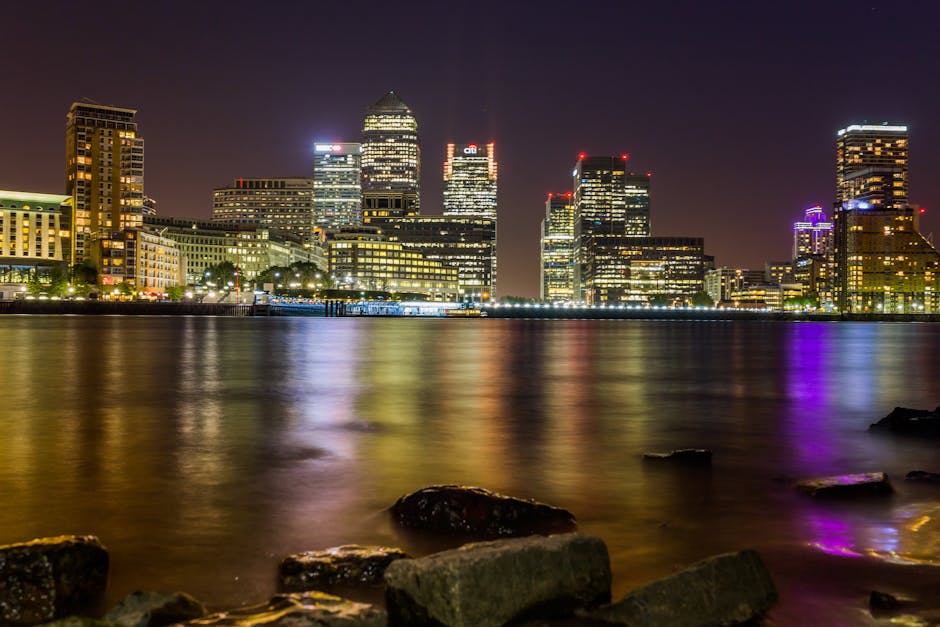 A nighttime cityscape of a modern financial district with illuminated high-rise buildings reflecting on the calm water of a river or harbour. The buildings display various architectural styles, with some featuring glass curtain walls and others with concrete or metal facades, all outlined with bright lights and signage. The skyline includes several buildings with prominent lighting accents, such as purple, red, and white lights, creating a vibrant atmosphere against the dark sky. In the foreground, there are dark, wet rocks along the shoreline, adding texture and depth to the scene. The water’s surface is smooth with slight ripples, mirroring the colorful city lights. The environment suggests an urban environment at night, with the scene captured from a vantage point on a waterfront promenade or rocky embankment. This image highlights the contrast between the lively, illuminated city skyline and the tranquil water, subtly reflecting the possibility of alternative waste handling options, such as private waste removal or on-site clearance, that complement traditional rubbish collection services.