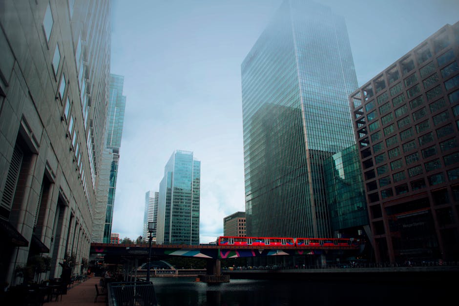 A cityscape featuring modern high-rise buildings with glass facades reflecting the surrounding skyline and sky. On the left, a beige stone building with rectangular windows and decorative architectural details is visible. In the background, several skyscrapers of varying heights, predominantly made of glass and steel, dominate the scene. A red train is traveling along an elevated track that runs across the lower part of the image, partially obscuring the waterway or canal beneath it. The scene is illuminated by natural daylight, with some haze or mist softening distant building outlines, creating a contemporary urban environment. The image aligns with the context of urban rubbish removal services, exemplifying areas where independent waste collection or on-site clearance could be necessary for maintaining city cleanliness, as highlighted by Rubbish Clearance Docklands providing tailored rubbish disposal solutions in such environments.