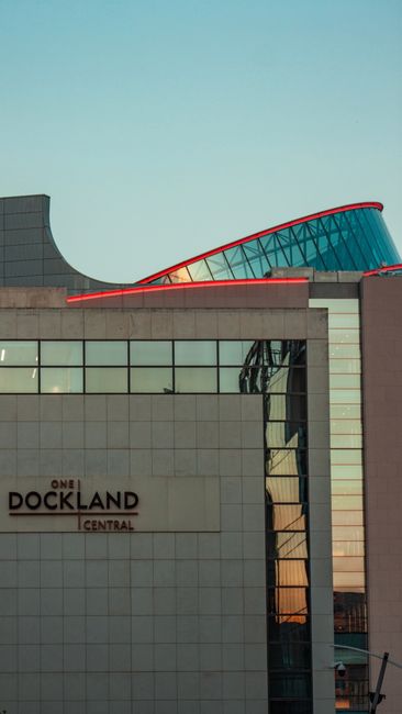 Close-up view of the upper section of a modern commercial building in Docklands, featuring a mix of materials including grey concrete, large glass windows with reflective surfaces, and a distinctive curved glass roof with a red edging. The building has a clean, structured façade with horizontal and vertical lines, and a visible sign reading 'ONE DOCKLAND CENTRAL' on the lower part of the exterior. The background shows a clear blue sky, highlighting the building's contemporary architectural design. This scene may relate to private or on-site waste management for commercial properties, as part of the broader context of rubbish removal and disposal services such as those provided by Rubbish Clearance Docklands.