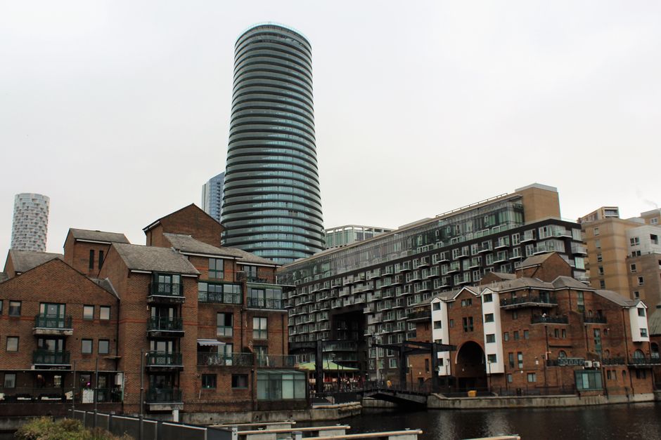 The image shows a cluster of modern residential buildings with various architectural styles and materials. In the foreground, there are several low-rise brick structures with pitched roofs, small balconies, and large windows, reflecting a traditional brick finish with a mix of red and brown tones. These buildings are situated along a narrow canal or waterway, with some small docks or jetties visible along the edge. Behind these, a large, contemporary multi-story apartment complex features extensive glass balcony railings, dark grey metal panels, and a layered, textured façade, creating a distinctive contrast with the brick buildings. In the background, a tall, cylindrical high-rise tower with horizontal black and white banding dominates the skyline, indicative of modern urban architecture. The scene is illuminated by natural daylight under an overcast sky, contributing to a neutral, professional appearance. This setting exemplifies a mixed residential environment that may be associated with alternative waste management approaches, such as private or on-site clearance services, aligned with the relevance of rubbish removal activities in urban development areas. Rubbish Clearance Docklands might undertake such clearance tasks in similar environments.