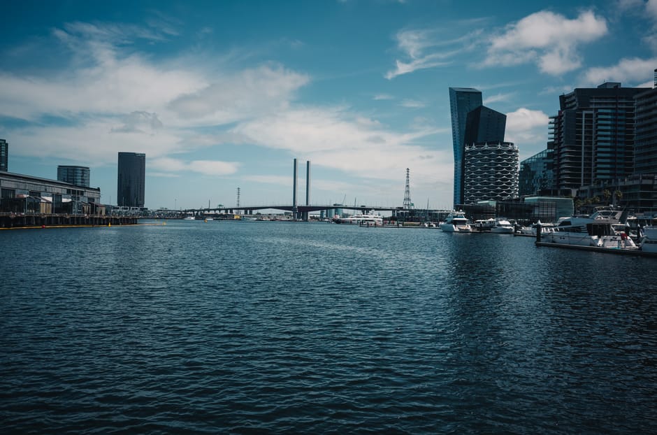 A wide view of a modern urban waterfront with a calm body of water in the foreground, dotted with several docked yachts and boats. On the left, there are industrial-style buildings with flat roofs and metallic finishes, possibly warehouses or commercial spaces, aligned along a paved quay. To the right, a cluster of tall, contemporary skyscrapers with varying geometric shapes, including one with a curved facade and another with an angular, glass-fronted design, dominate the skyline. In the background, a bridge with prominent support pillars spans across the water, connecting different parts of the city, and a transmission tower rises behind it. The sky is partly cloudy with patches of blue, and soft natural light illuminates the scene, capturing the clean, professional environment typical of a central business district near the water. This urban landscape exemplifies the proximity of commercial and residential buildings to waterfront leisure facilities, aligning with the context of independent waste management and rubbish removal services in a city setting, as offered by Rubbish Clearance Docklands.