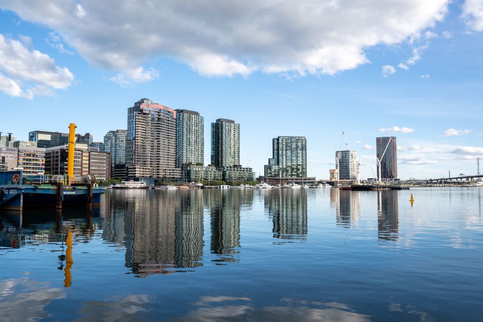 A wide view of a city skyline across a calm body of water during daytime features modern high-rise buildings with glass facades reflecting the sky. Several of these buildings are rectangular with varying heights, some with balconies and distinct architectural designs. In the foreground, part of a blue and black barge or boat, with visible metallic textures, is docked on the water's edge on the left side of the image, supported by wooden pilings painted yellow. The water surface is smooth with gentle ripples, creating clear reflections of the buildings and sky, which is partly cloudy with a mix of white clouds and blue sky. In the background, there is a bridge extending from right to left, and a construction crane is visible amongst the buildings, indicating ongoing development. This scene illustrates an urban waterfront environment where private land or water-based cargo handling may supplement traditional rubbish removal services, which could be relevant to discussions on skip alternatives in docklands rubbish removal by Rubbish Clearance Docklands.