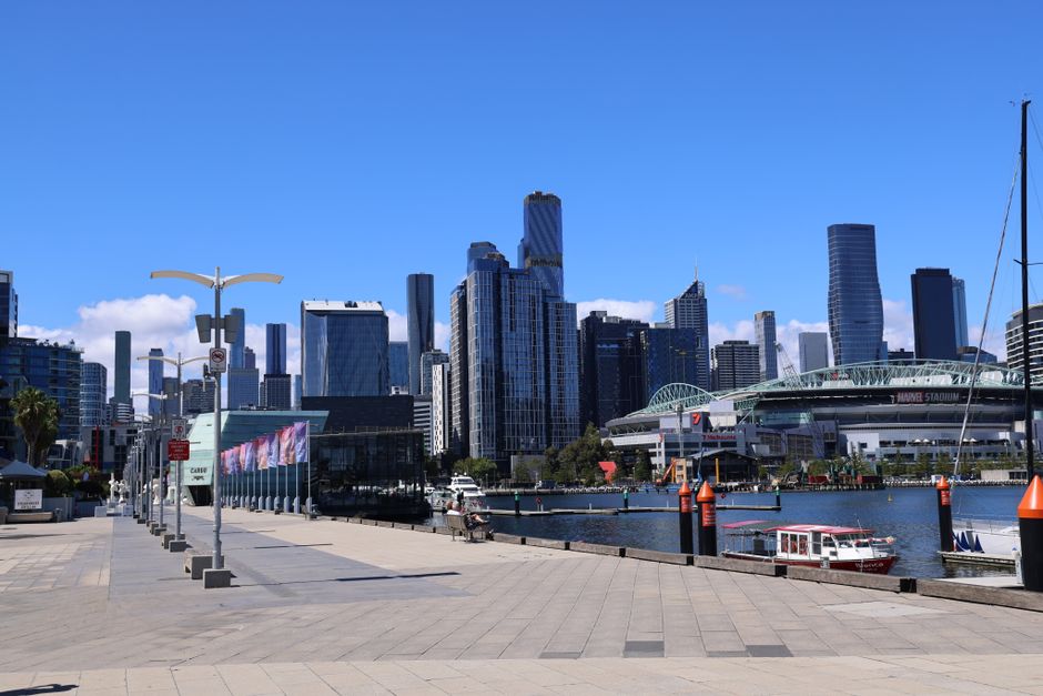 A wide view of a modern urban waterfront with a calm body of water in the foreground, dotted with several docked yachts and boats. On the left, there are industrial-style buildings with flat roofs and metallic finishes, possibly warehouses or commercial spaces, aligned along a paved quay. To the right, a cluster of tall, contemporary skyscrapers with varying geometric shapes, including one with a curved facade and another with an angular, glass-fronted design, dominate the skyline. In the background, a bridge with prominent support pillars spans across the water, connecting different parts of the city, and a transmission tower rises behind it. The sky is partly cloudy with patches of blue, and soft natural light illuminates the scene, capturing the clean, professional environment typical of a central business district near the water. This urban landscape exemplifies the proximity of commercial and residential buildings to waterfront leisure facilities, aligning with the context of independent waste management and rubbish removal services in a city setting, as offered by Rubbish Clearance Docklands.